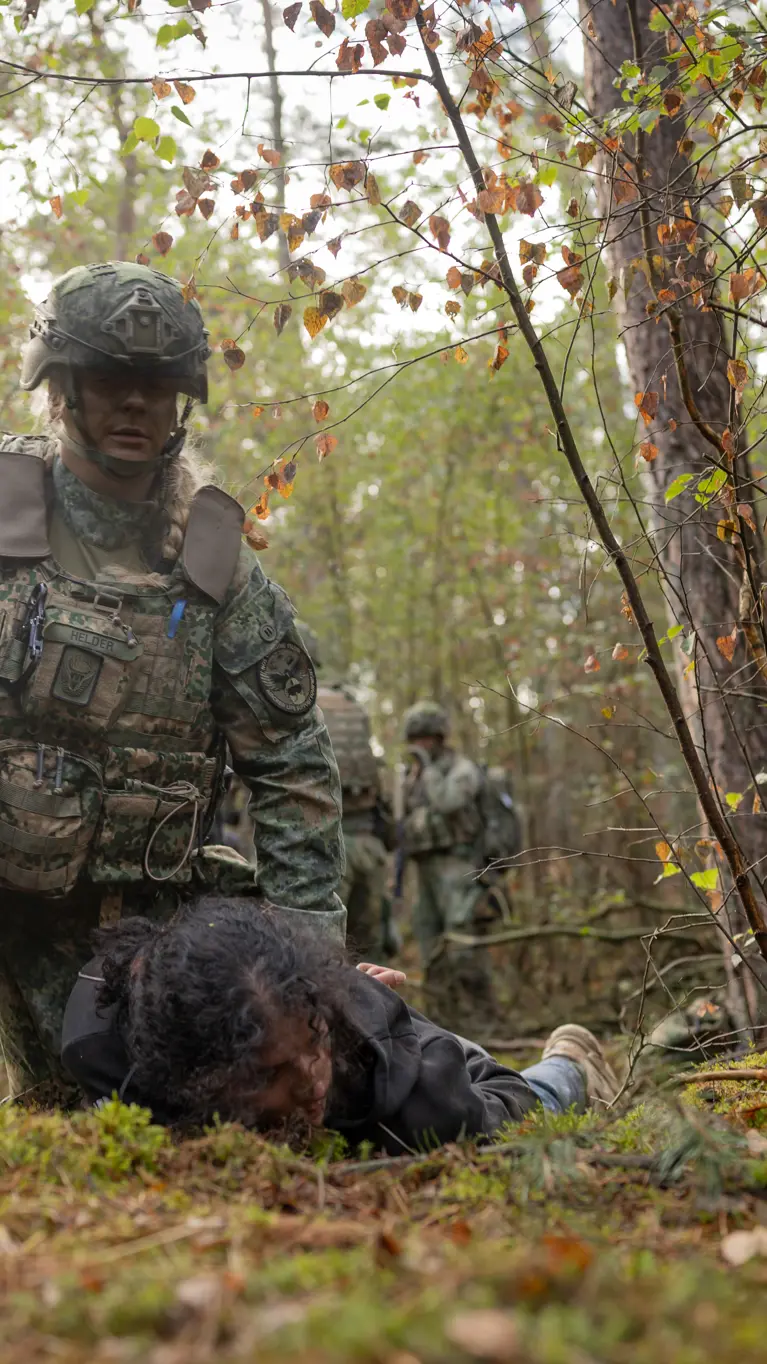Twee militairen in het bos houden ieder een persoon in burgerkleding tegen de grond gedrukt.