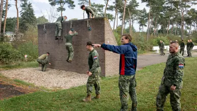 Een sportinstructeur wijst, terwijl achter hem militairen over een muurtje klimmen. 