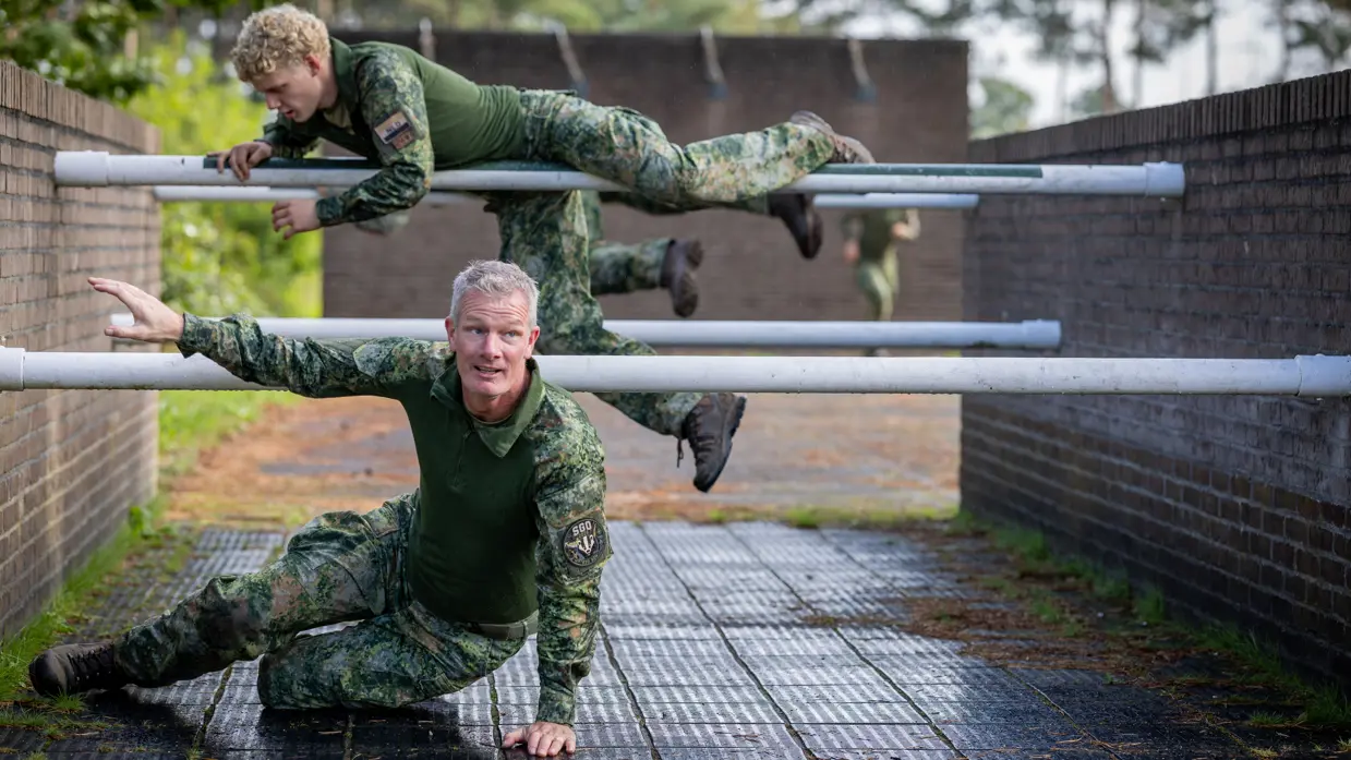 Twee militairen bewegen zich over en onder een obstakel met horizontale palen door. 
