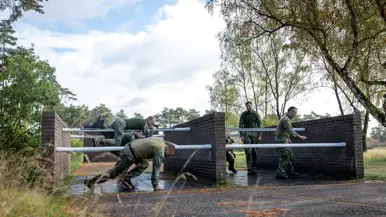 Een sportinstructeur wijst, terwijl achter hem militairen over een muurtje klimmen. Twee militairen bewegen zich over en onder een obstakel met horizontale palen door. Een militair springt over een obstakel op de hindernisbaan. Twee militairen kruipen door een obstakel op de hindernisbaan.
