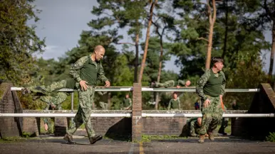 Een sportinstructeur wijst, terwijl achter hem militairen over een muurtje klimmen. Twee militairen bewegen zich over en onder een obstakel met horizontale palen door. Een militair springt over een obstakel op de hindernisbaan. Twee militairen kruipen door een obstakel op de hindernisbaan.