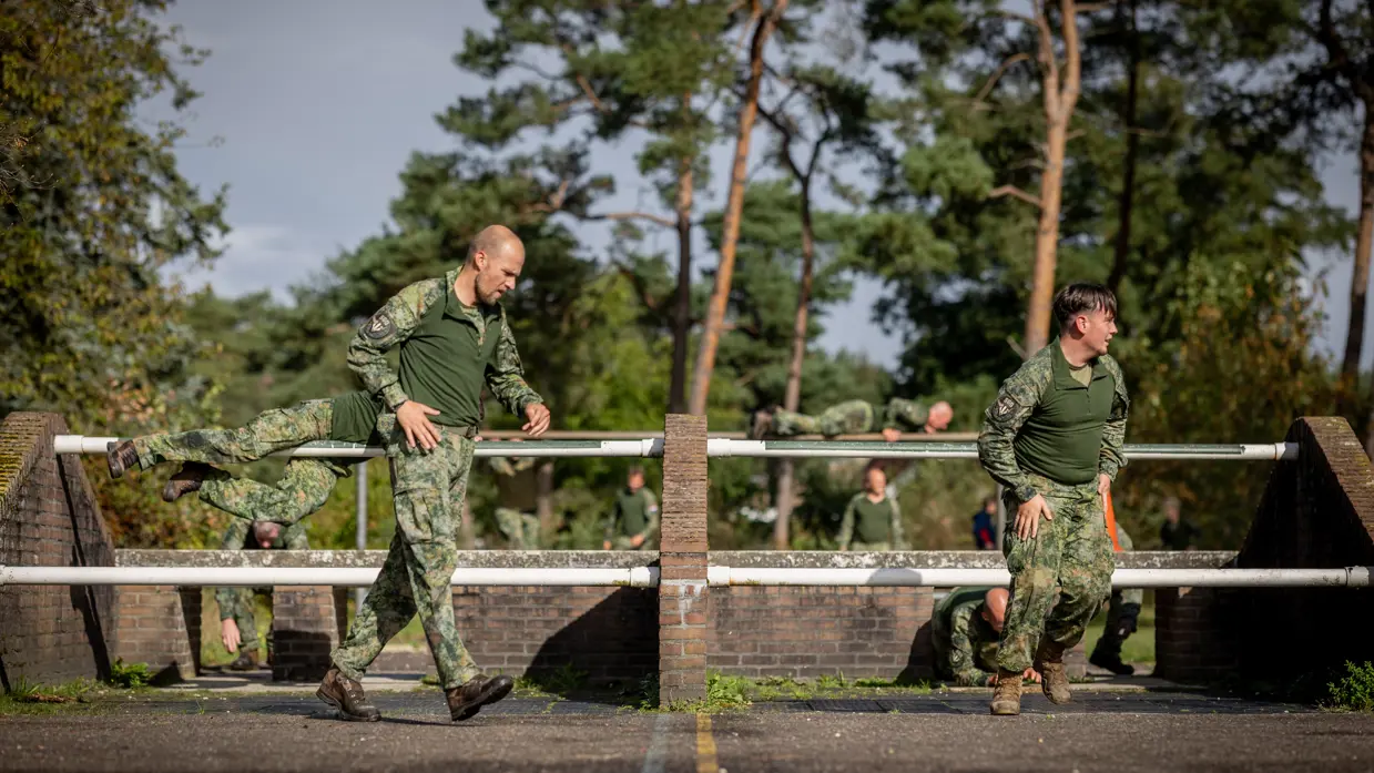 Een sportinstructeur wijst, terwijl achter hem militairen over een muurtje klimmen. Twee militairen bewegen zich over en onder een obstakel met horizontale palen door. Een militair springt over een obstakel op de hindernisbaan. Twee militairen kruipen door een obstakel op de hindernisbaan.