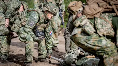 Een groepje militairen tilt een brancard van de grond op een zanderige weg. De militairen lopen met de brancard.