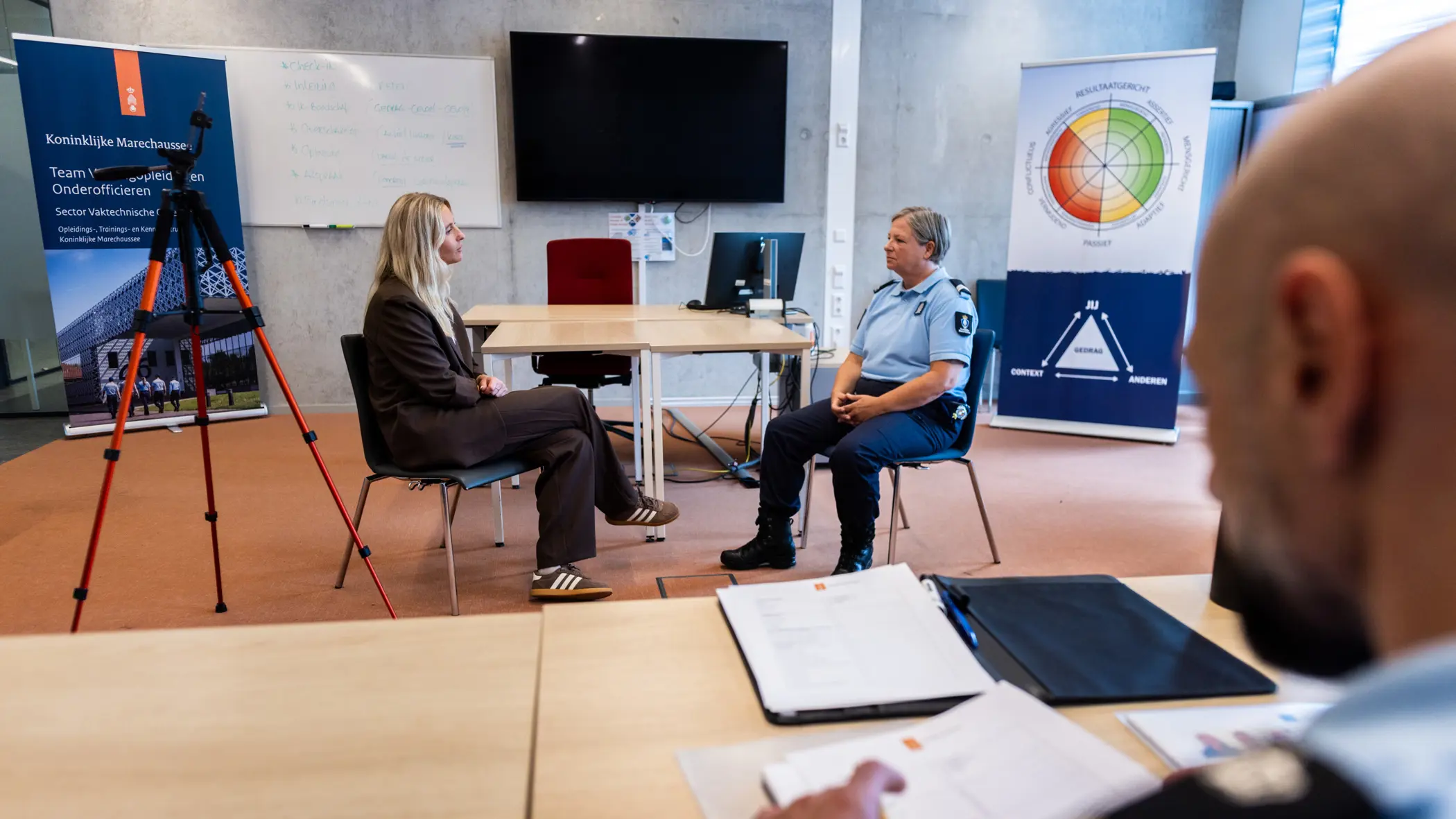 Een acteur en marechaussee in gesprek, de instructeur kijkt toe.