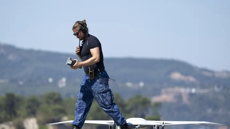 Een man met donkerblauw T-shirt en blauwe broek loopt met een portofoon in zijn handen weg van een grijze drone. 