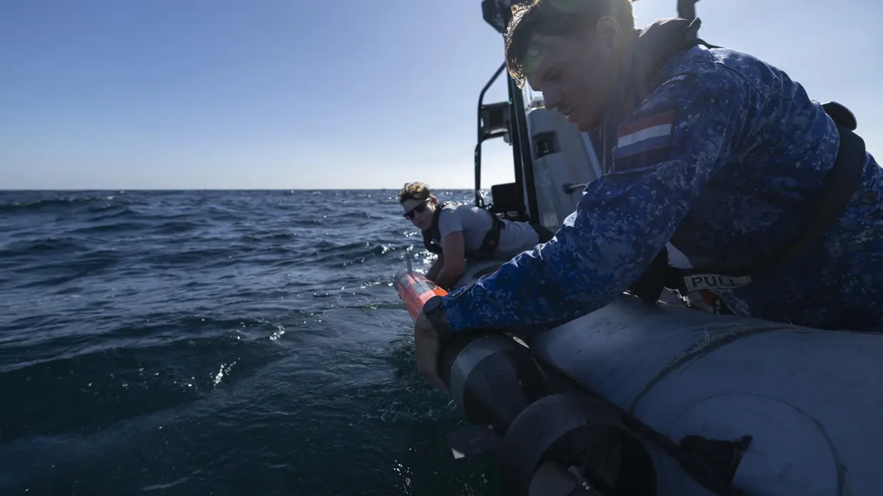 Een militair van de marine en een vrouw met grijs t-shirt leggen gezamenlijk een oranje drone in het water vanaf een bootje.