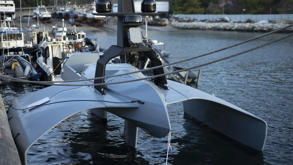 Een grijs futuristisch bootje ligt aan de kade. Op de achtergrond vissersbootjes. 