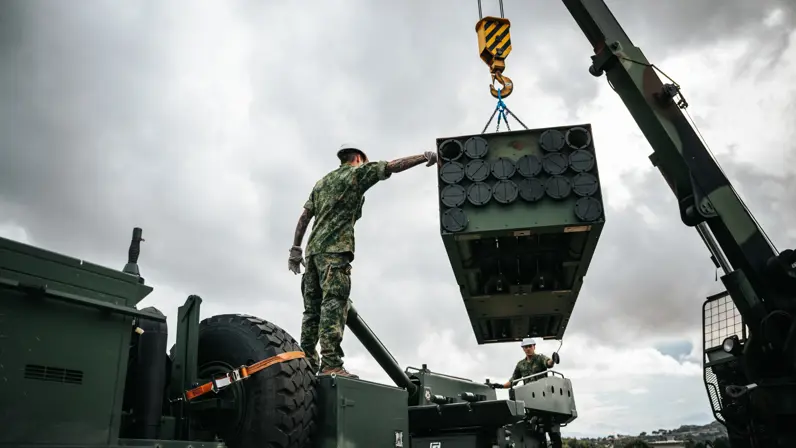 Een hijskraan tilt een soort container in de lucht, dit gebeurt onder begeleiding van twee militairen met een witte helm op.