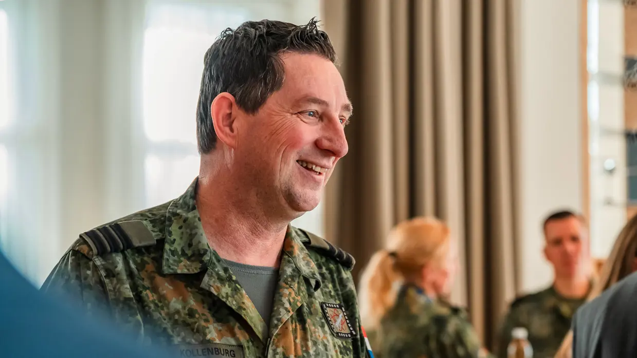 Een Nederlandse militair kijkt lachend naar rechts, op de achtergrond een paar militairen in gesprek.