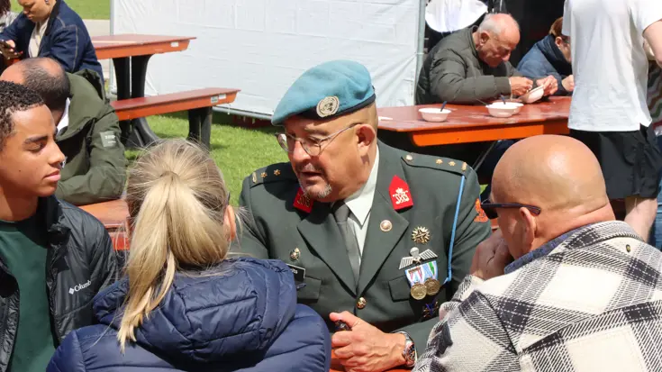 Een militair in uniform met een blauwe baret zit aan een tafel. Hij is in gesprek met jongeren. 