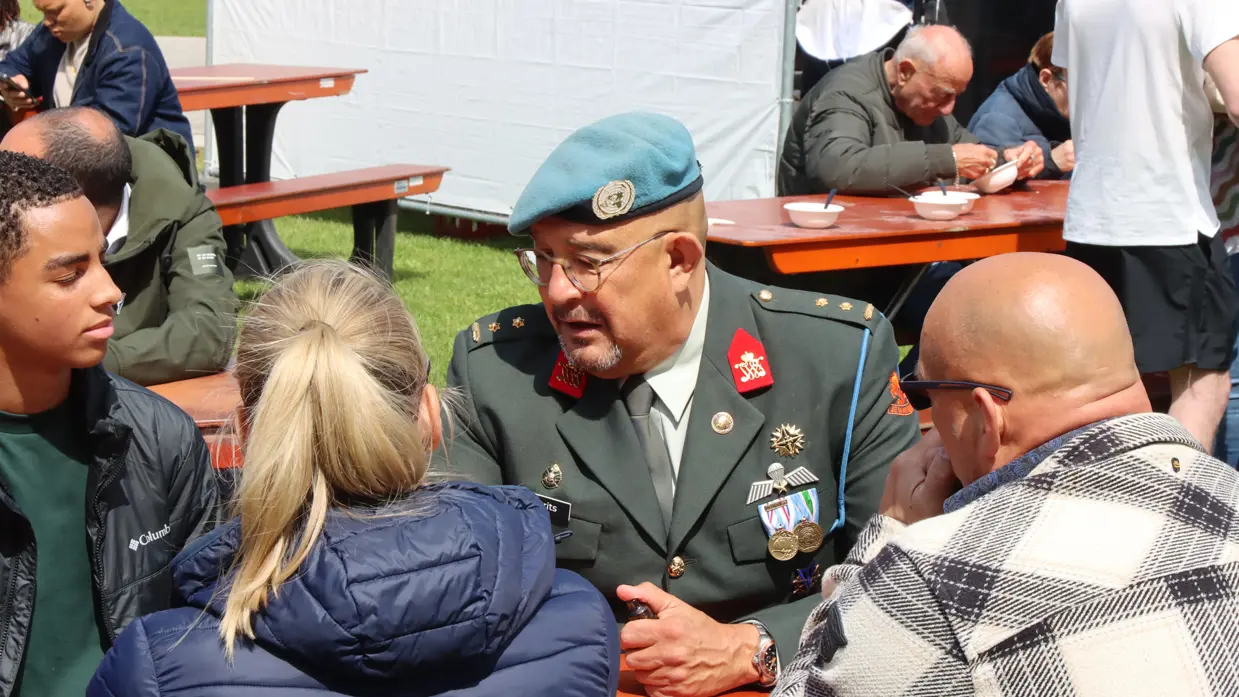 Een militair in uniform met een blauwe baret zit aan een tafel. Hij is in gesprek met jongeren. 