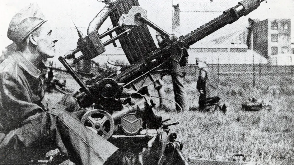 Een soldaat in uniform bedient een zware, oude machinegeweeropstelling in een open veld met gras en gebouwen op de achtergrond. 
