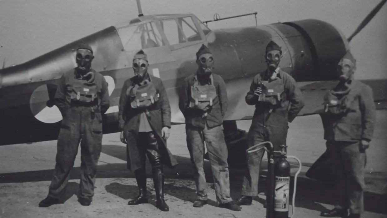 Vijf mannen in vliegtenue met gasmaskers en veldmasker staan voor een oud militair vliegtuig, met een brandblusapparaat naast hen. De foto is zwart-wit.