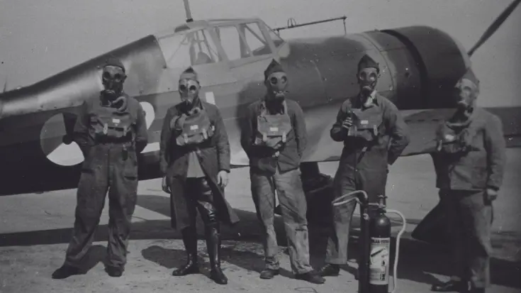 Vijf mannen in vliegtenue met gasmaskers en veldmasker staan voor een oud militair vliegtuig, met een brandblusapparaat naast hen. De foto is zwart-wit.