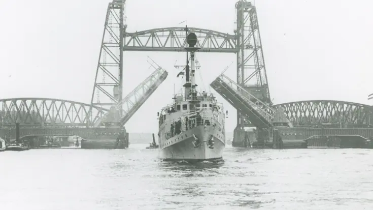 Een groot schip vaart onder een hoge, metalen ophaalbrug door. De brug is open en heeft twee torens met een roosterachtige constructie boven het water.