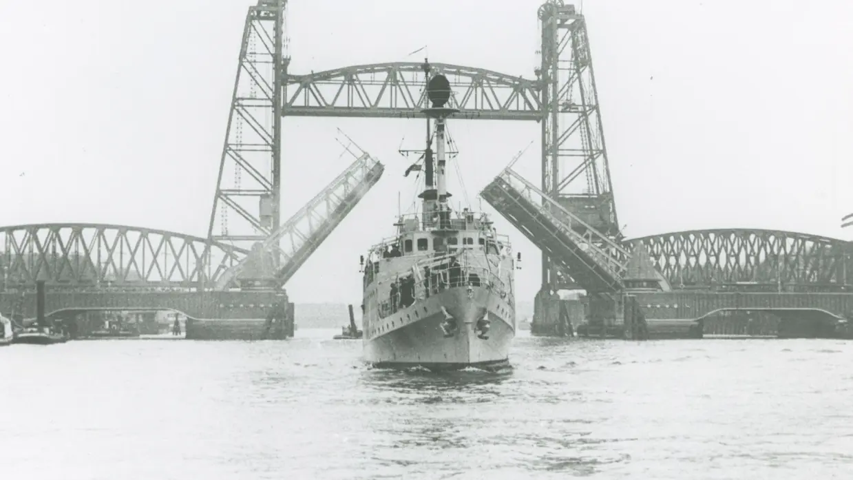 Een groot schip vaart onder een hoge, metalen ophaalbrug door. De brug is open en heeft twee torens met een roosterachtige constructie boven het water.
