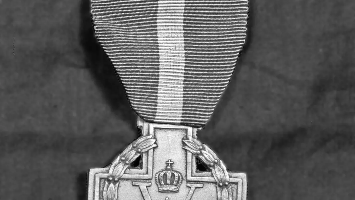 Links: Een vrouw in een dikke jas met bontkraag is omringd door mannen in uniformen in een haven. Rechts: Een metalen medaille met een krans, een "W" in het midden, en een kroontje erboven, aan een verticaal gestreept lint.
