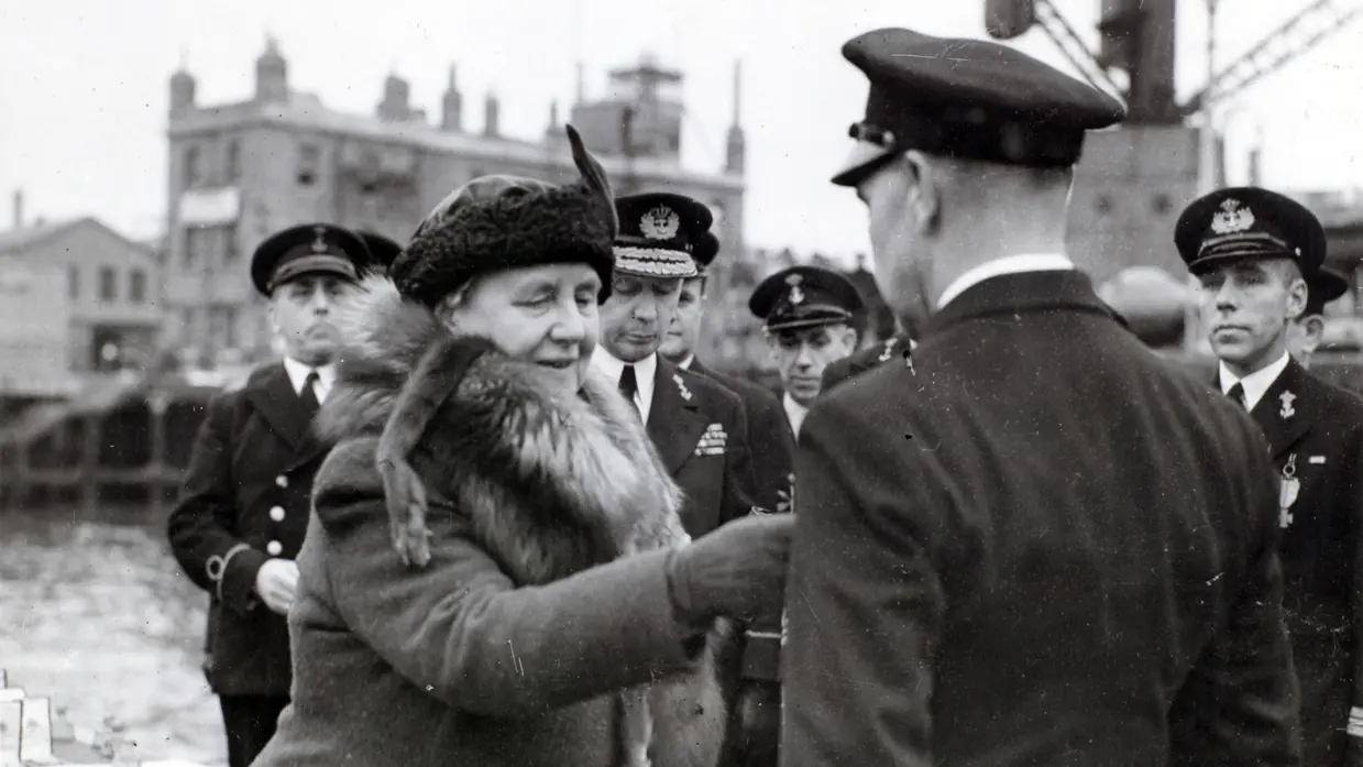 Links: Een vrouw in een dikke jas met bontkraag is omringd door mannen in uniformen in een haven. Rechts: Een metalen medaille met een krans, een "W" in het midden, en een kroontje erboven, aan een verticaal gestreept lint.