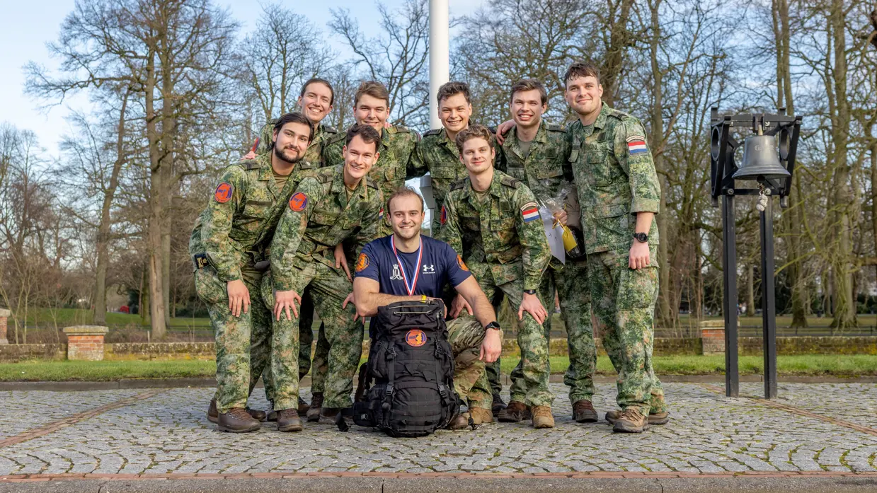 Zeven mannen in militaire kleding staan glimlachend in een groep buiten, met bomen op de achtergrond. Een man knielt voor hen met een grote rugzak en een medaille om. 