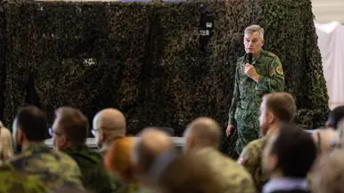  Links: Een militair in uniform staat voor een groen gecamoufleerd doek en spreekt met een microfoon. Een groep mensen in uniform en burgerkleding luistert aandachtig. Rechts: Een bord met het embleem "Casualty Move 2026 Medical Quadrigga 2026" staat op de voorgrond. Achterin zitten mensen in een grote ruimte naar een presentatie te kijken.