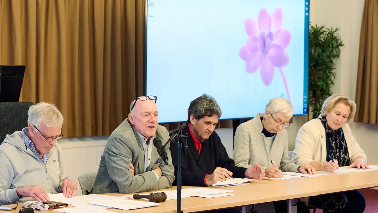 Statige heren en dames zitten achter een lange tafel, waarvan een achter een microfoon.&nbsp;