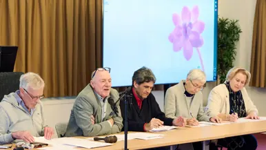 Statige heren en dames zitten achter een lange tafel, waarvan een achter een microfoon.&nbsp;
