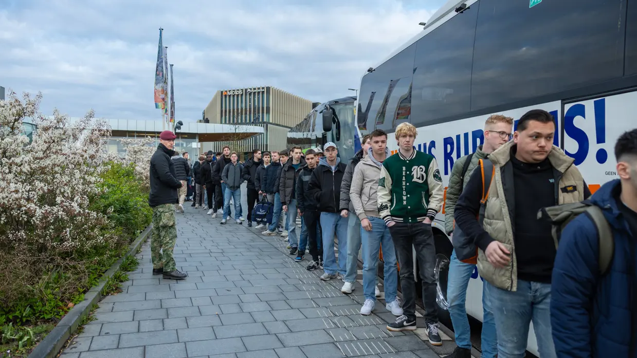 Een groep jongeren staat buiten in een rij voor een bus met "Rubens!" erop. Sommigen dragen jassen en rugzakken, een militair staat opzij. 
