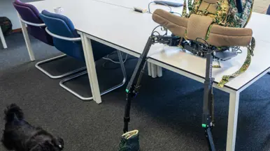 Een exoskelet met camouflage rugzak en groene bergschoenen staat op een tafel in een klaslokaal met stoelen en tafels.