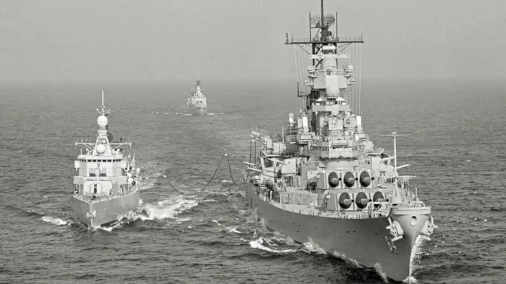  Drie oorlogsschepen varen op ruwe zee: twee grote schepen voorop, verbonden door kabels, en een kleiner schip op de achtergrond.