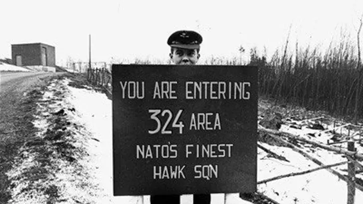 Militair langs weg voor bord met Engelstalige tekst: you are entering 324 area natos finest hawk sqn.