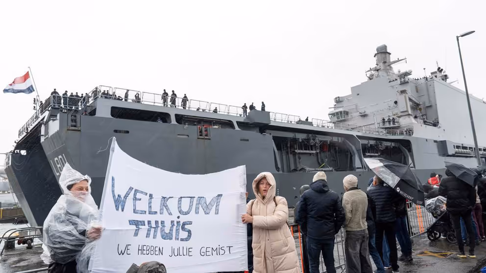 Mensen in regenkleding met spandoek Welkom Thuis bij aankomst marineschip.