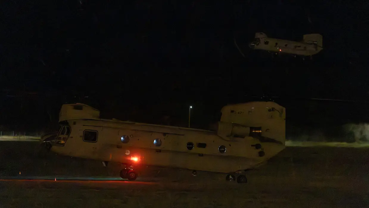 Twee Chinooks in het duister. Een in de lucht, de ander komt net los van het grasveld.