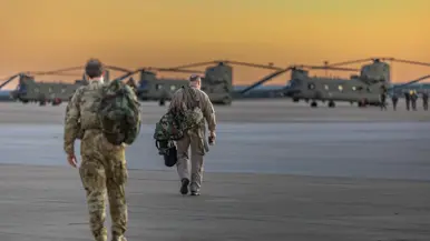 Twee militairen lopen met tassen richting drie Chinooks op een platform. Op de achtergrond een oranje-gele lucht.