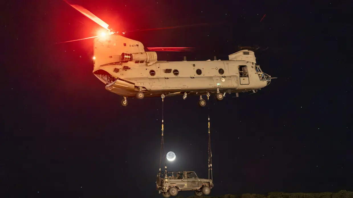 Een bij duisternis wegvliegende Chinook met eronder een militair voertuig aan kabels. Eronder begroeiing. Tussen de helikopter en het voertuig is de maan te zien.