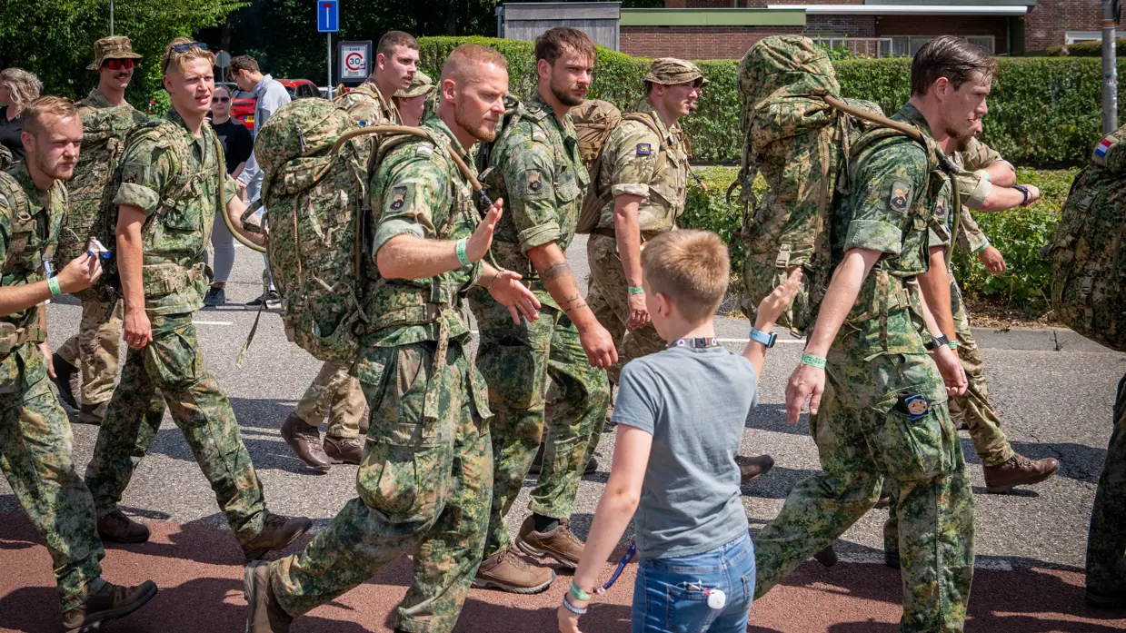 Een groep militairen marcheert door een straat. Een militair high fivet met een jongetje langs de weg.  