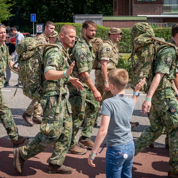 Een groep militairen marcheert door een straat. Een militair high fivet met een jongetje langs de weg.  