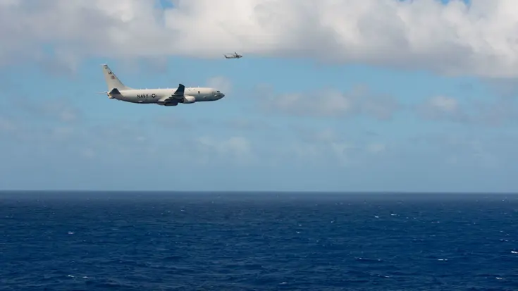 Een groot militair vliegtuig vliegt met de zee onder en blauwe lucht boven. 