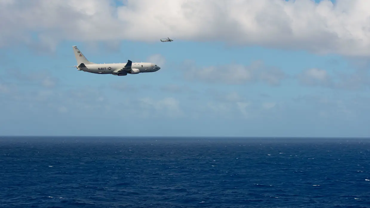 Een groot militair vliegtuig vliegt met de zee onder en blauwe lucht boven. 