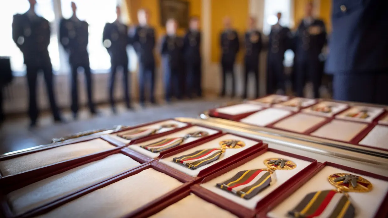 Medailles in doosjes met op de achtergrond onscherp militairen in dagelijks tenue. 