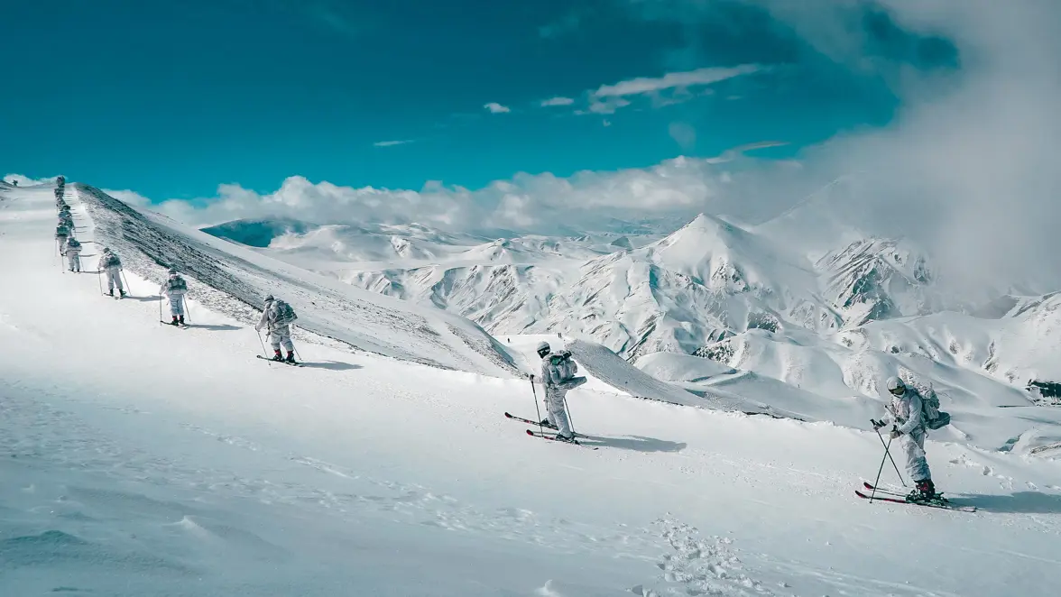 Vijf mensen in witte winterkleding skiën in een rij over een besneeuwde berghelling met indrukwekkende, besneeuwde bergen en een blauwe lucht op de achtergrond. 