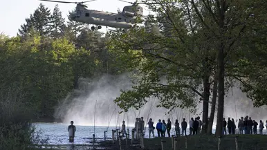 Helikopter schept water op uit een meer, groene bomen, toeschouwers kijken toe vanaf de oever in avondlicht. 