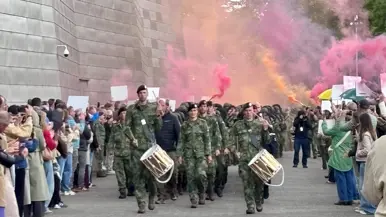 Militairen lopen door een haag van vrienden en familie en rode en oranje rookfakkels.