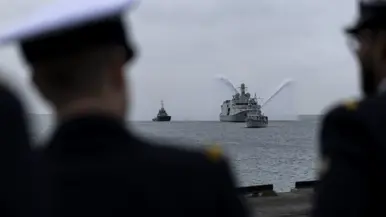 Marineschip de ‘Vlissingen’ op zee, daarnaast twee sleepboten die met hun waterkanonnen spuiten. Het schip wordt voorgegaan door een kleine mijnenveger. Op de tweede afbeelding hetzelfde schip, maar dan gezien langs een tweetal marineofficieren aan de wal. 
