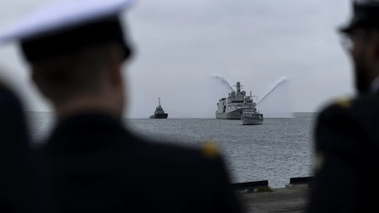 Marineschip de ‘Vlissingen’ op zee, daarnaast twee sleepboten die met hun waterkanonnen spuiten. Het schip wordt voorgegaan door een kleine mijnenveger. Op de tweede afbeelding hetzelfde schip, maar dan gezien langs een tweetal marineofficieren aan de wal. 