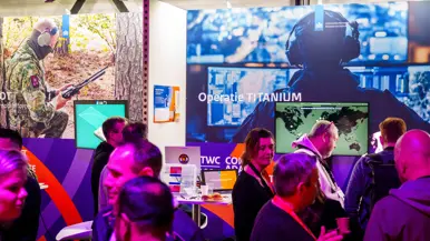 Groep mensen staat rond stand op conferentievloer. Een man wijst op computerscherm in een stand op de conferentie en een man in blauwe trui op de rug gezien zit tussen 2 mannen met VR-bril op.