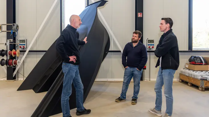 Drie mensen in fabriekshal in gesprek bij een onderdeel van 3D-geprinte boot.
