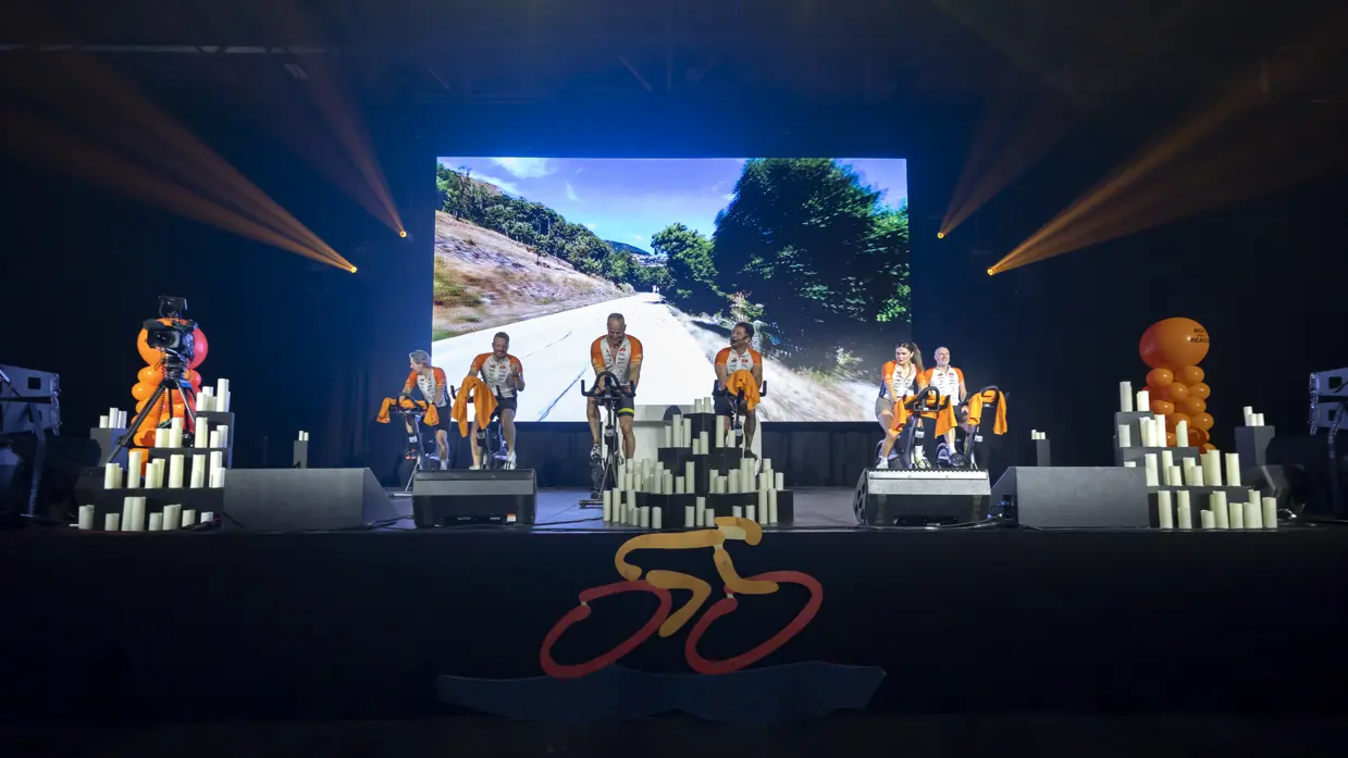 Deelnemers van team Marechaussee aan de Alpe d’HuZes Indoorride 2026 op een podium met beeldscherm erachter. 