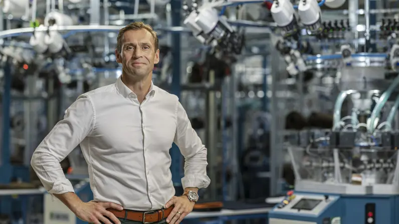 Een man in witte blouse poseert met zijn handen in de zij in een fabriekshal. Op de achtergrond staan grote machines.