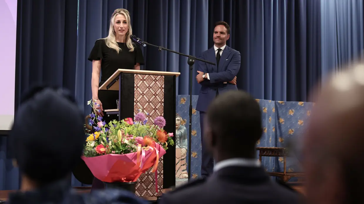  Amber, in zwarte jurk, staat achter een microfoon en spreekt de aanwezigen toe.