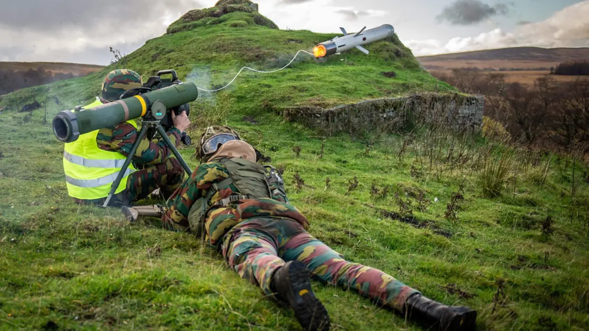 Een militair zit op de grond en vuurt vanaf zijn schouder een raket af. Deze komt met een dun touw erachter uit de afvuureenheid. Daarnaast ligt een militair op het grasveld. De twee volgen de baan van het projectiel.